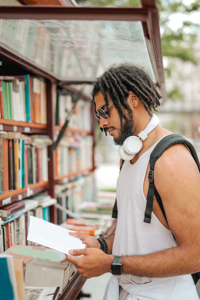 Confident adult Hispanic male in sunglasses and casual clothes with backpack and headphones reading book near bookshelf while making choice in stall on street